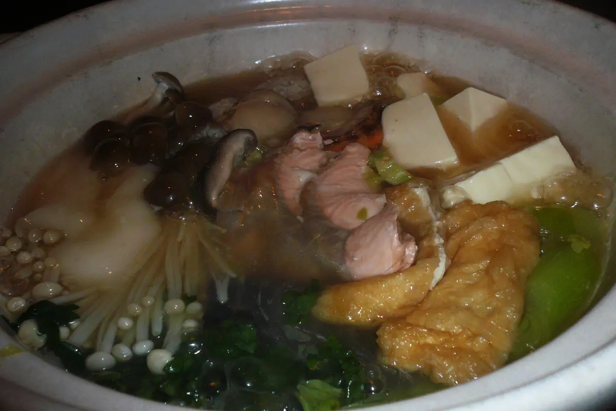 Una olla de barro con estofado chankonabe hirviendo sobre una mesa, con pollo en lonchas, tofu, setas, verduras de hoja y fideos en el caldo