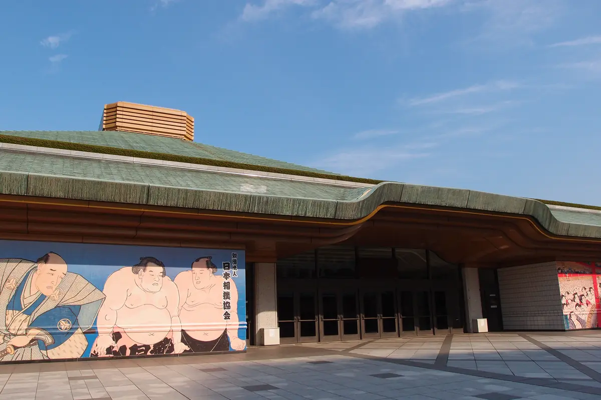 Dlouhá zelená střecha a vstupní hala sumo arény Ryogoku Kokugikan v Tokiu, s malovanou nástěnnou malbou zápasníků na zdi vedle dveří