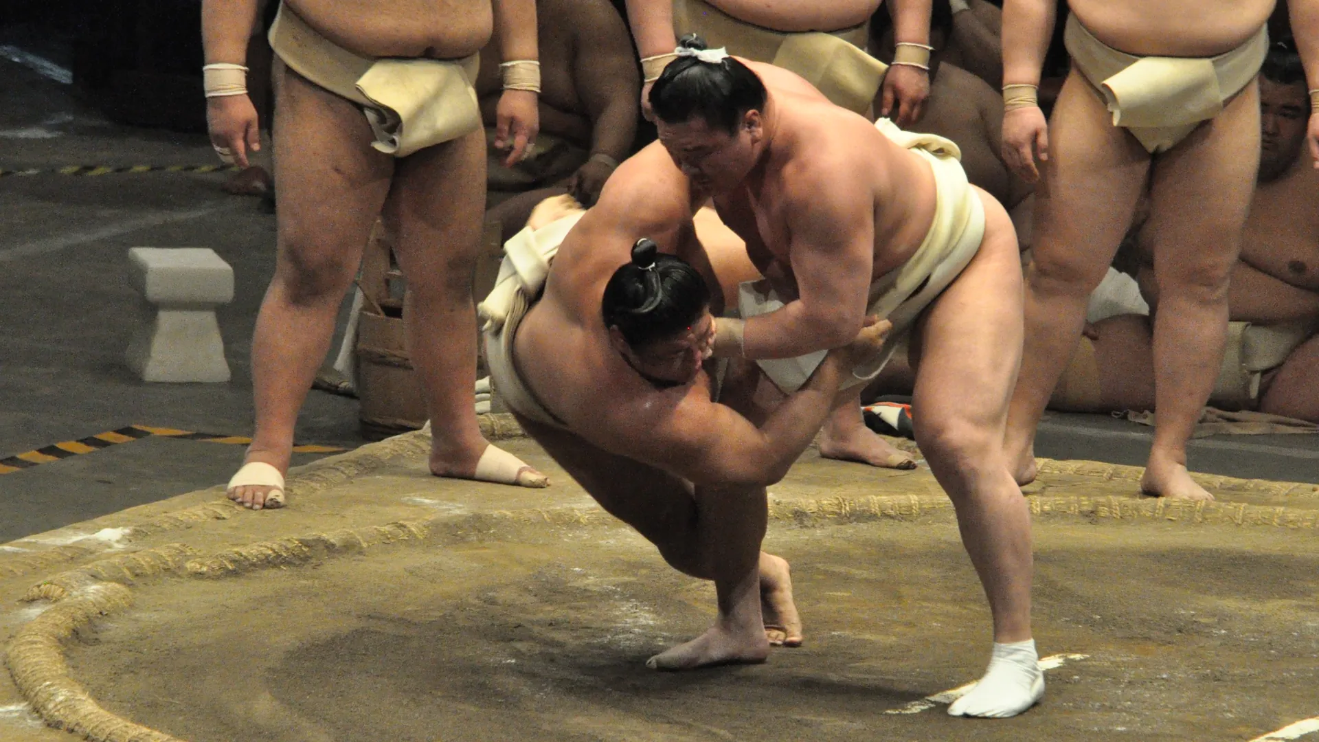 Dwóch zapaśników sumo w zwarciu na glinianym ringu obłożonym balami słomy ryżowej, a za nimi stoją i klęczą inni zapaśnicy