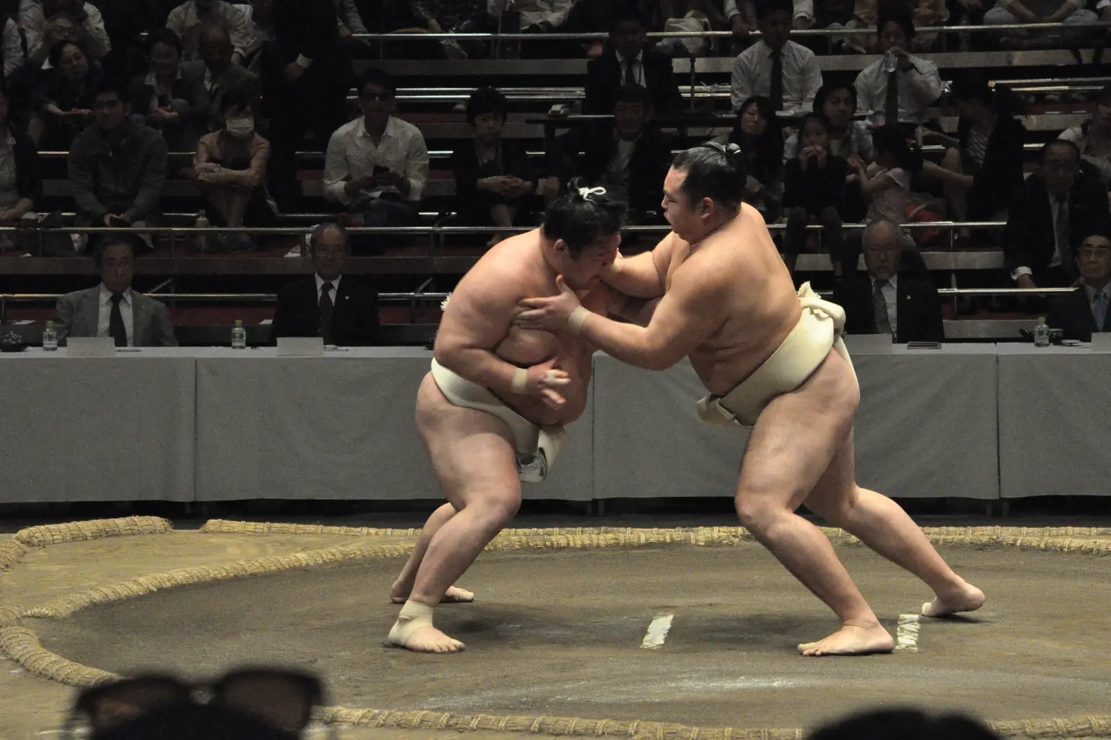 Due lottatori di sumo che si afferrano su un ring di argilla durante una sessione di allenamento, con il pubblico sugli spalti dietro e il tavolo dei giudici oltre il ring