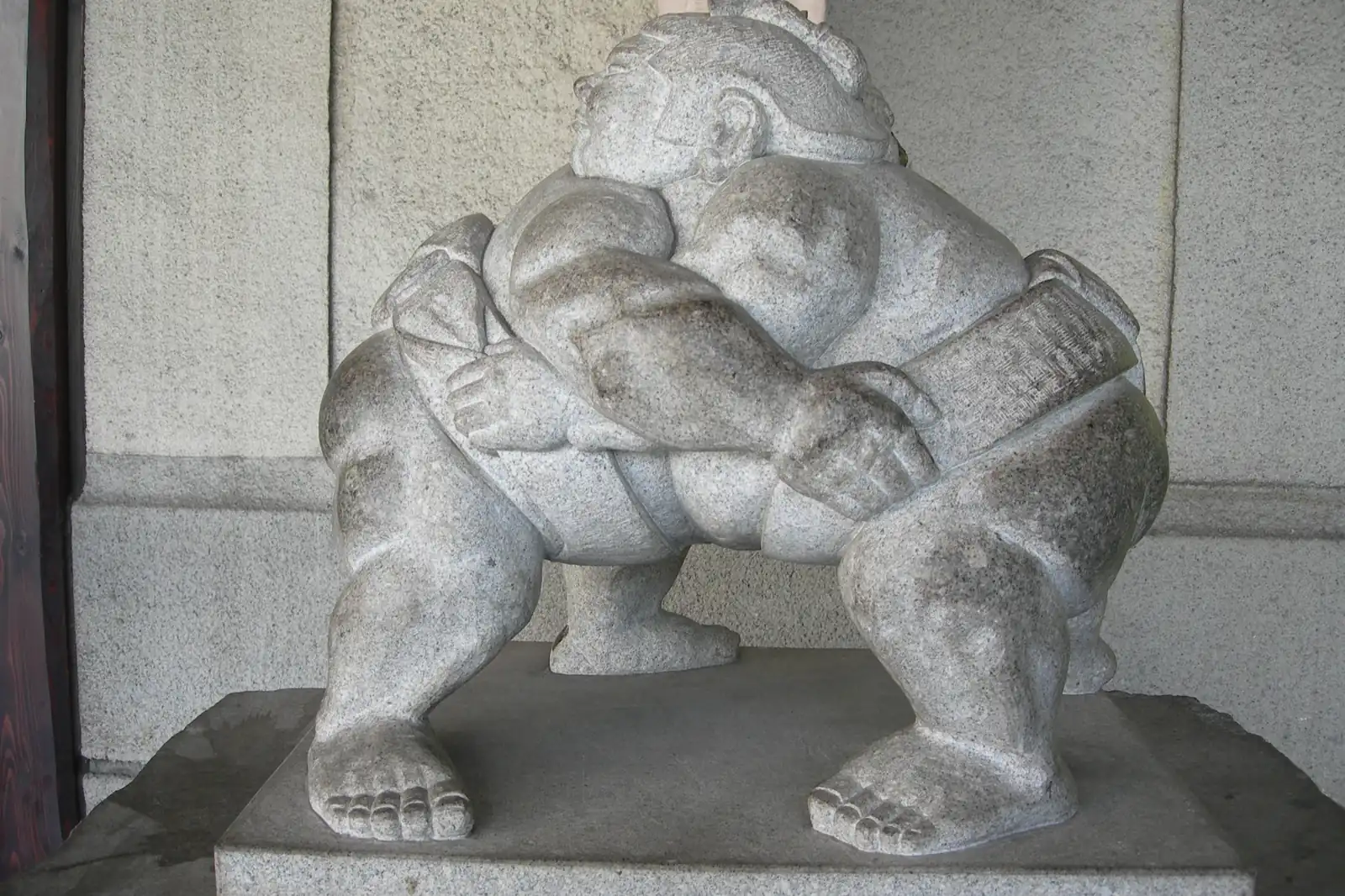 Una statua in pietra grigia di due lottatori di sumo avvinghiati, collocata all'ingresso di una stazione con una parete in pietra levigata sullo sfondo